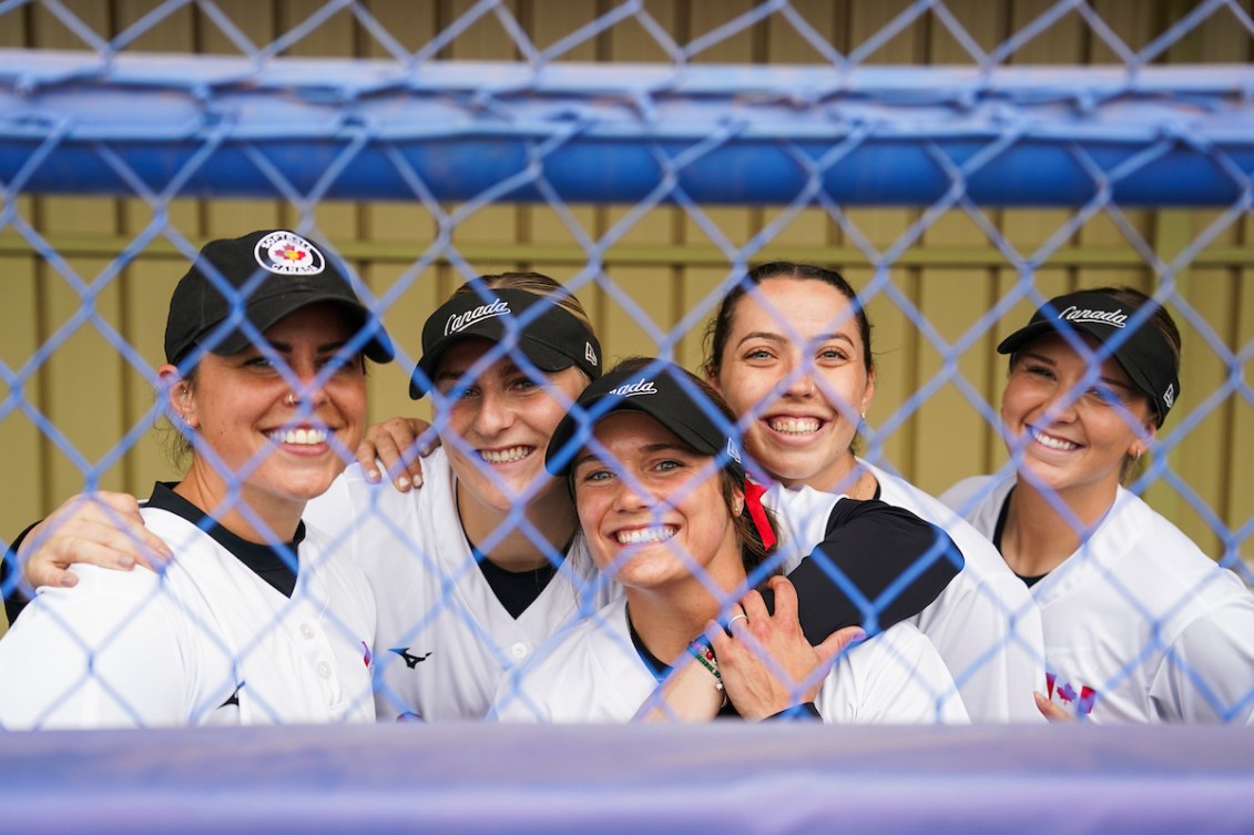 Team Canada softball players smile through the fence into the dugout