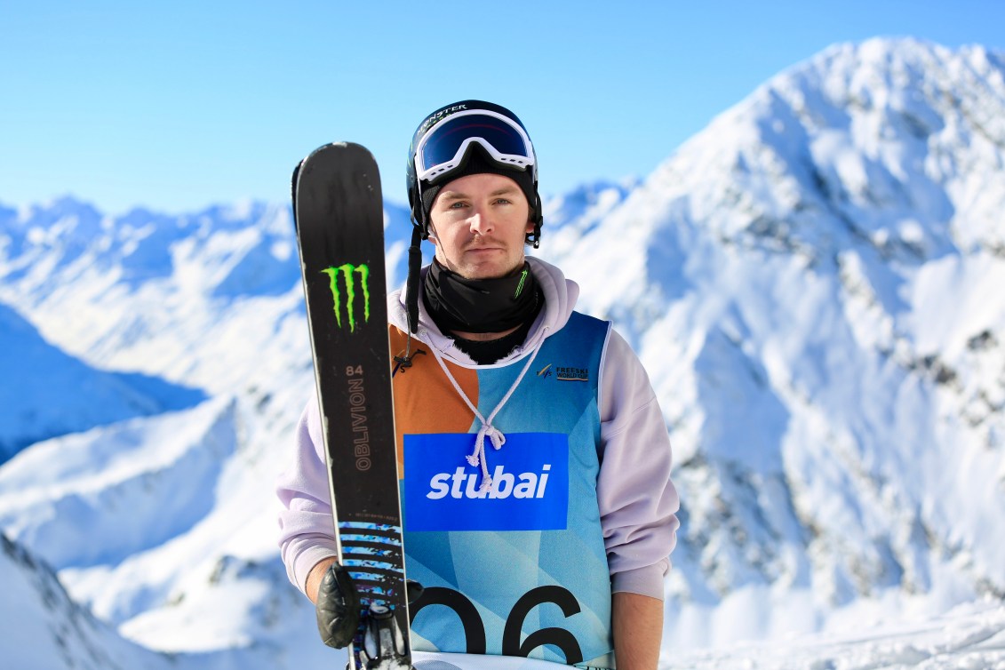 Evan McEachran poses for the camera in front of a mountain holding his skis by his side