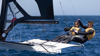 Two sailors in yellow life jackets hang off the side of their boat during a race
