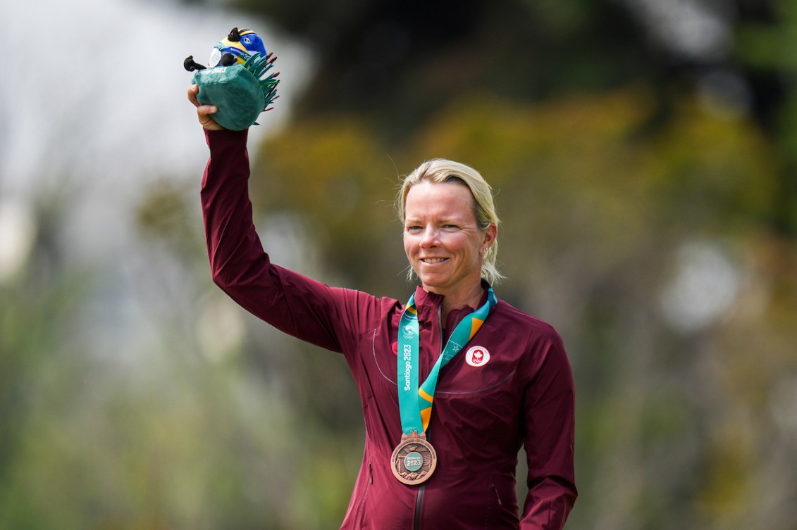 A female Team Canada athlete wears a bronze medal and holds a mascot stuffed animal, wearing Team Canada attire