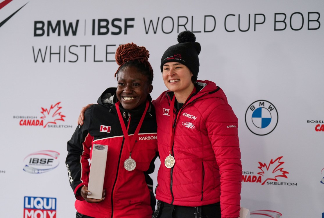 Cynthia Appiah and Bianca Ribi wear silve and gold medals while standing in front of an IBSF World Cup sign 