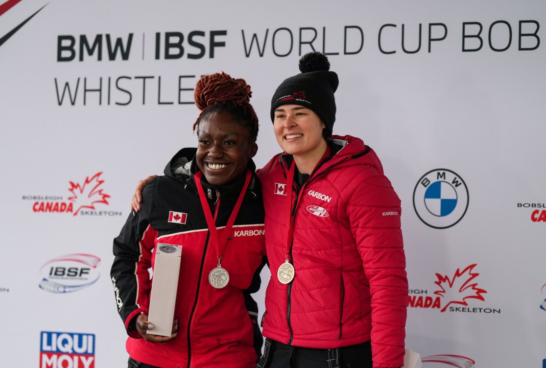 Cynthia Appiah and Bianca Ribi wear silve and gold medals while standing in front of an IBSF World Cup sign