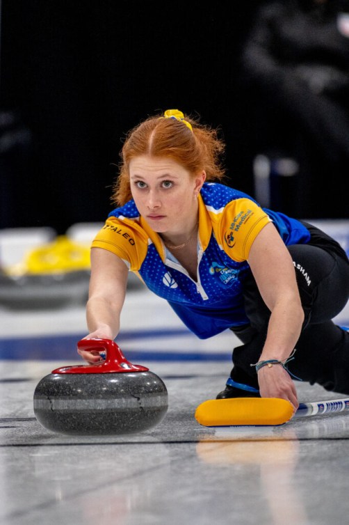 Curler in blue and yellow shirt slides on the ice to throw a red rock