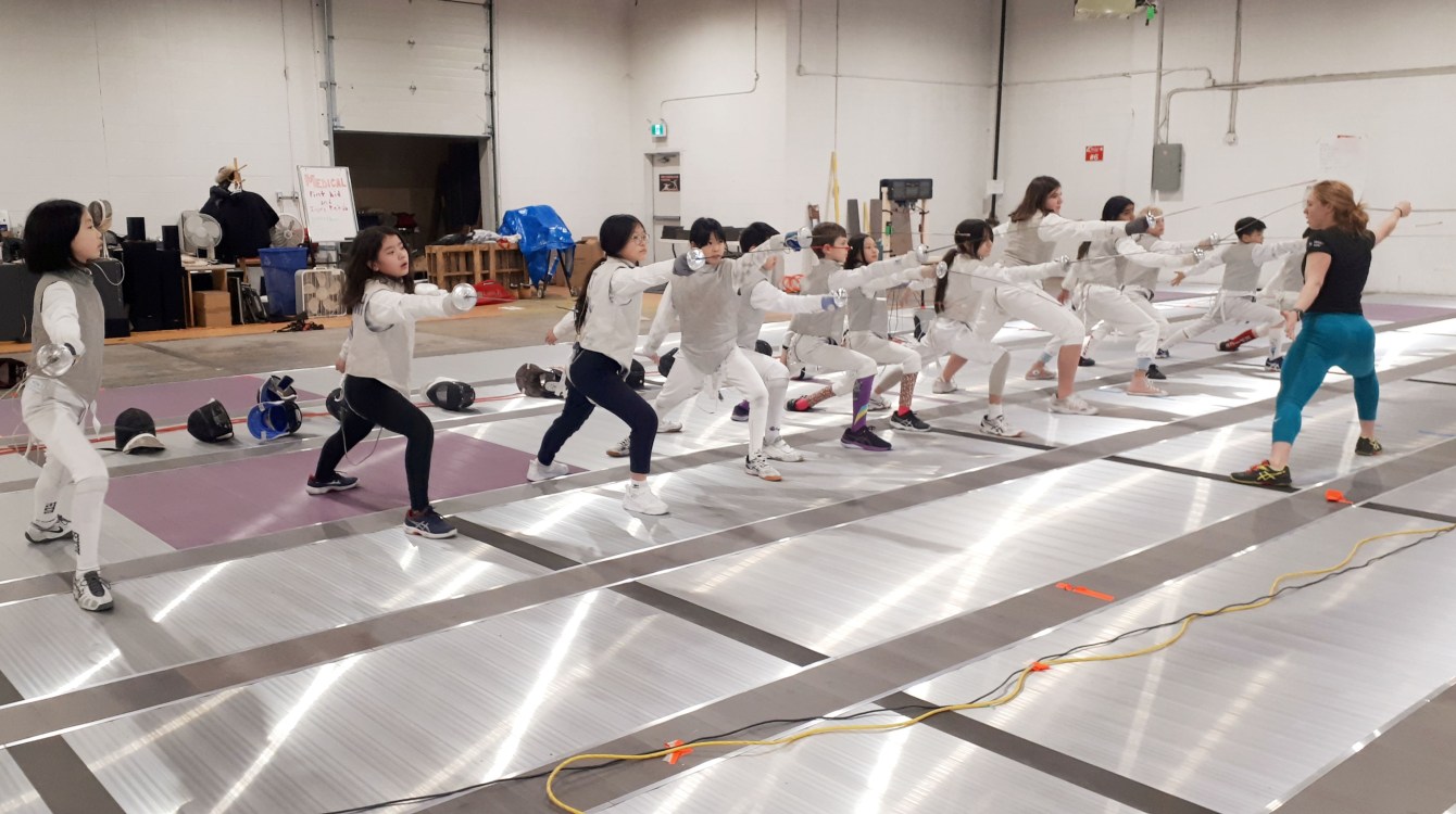 A group of young fencers stand in a line and practice their swordwork