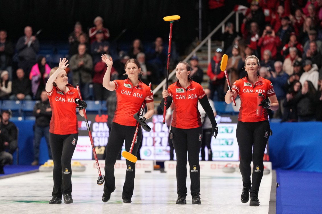 Four women curlers in red Canada uniforms hold their brooms up as they wave to the crowd