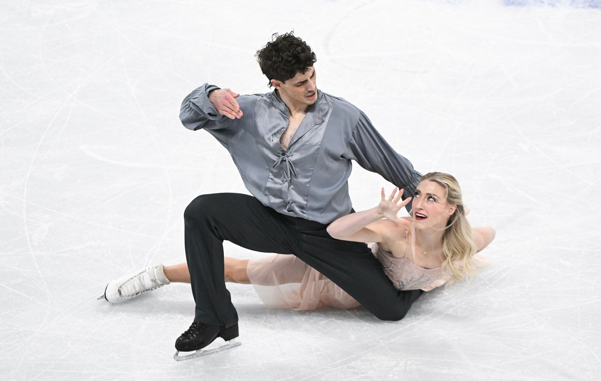 Piper Gilles in a light pink dress lies on the ice in a pose as she looks up at partner Paul Poirier in a grey shirt