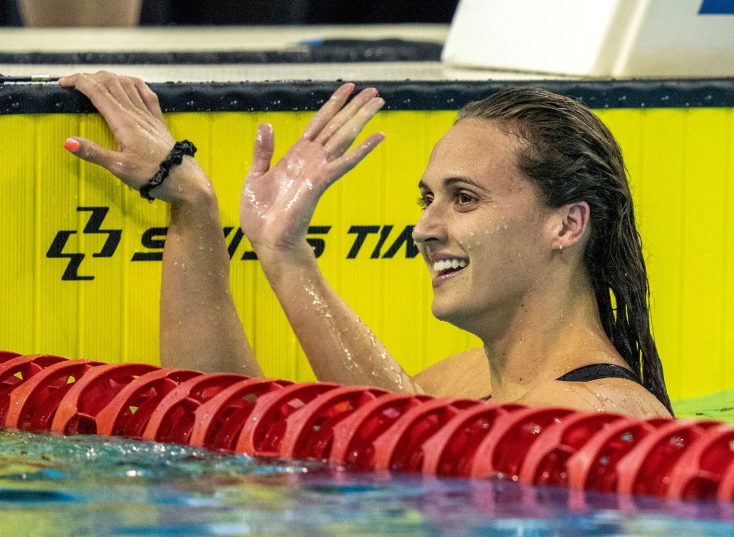 Kylie Masse waves as she holds onto the edge of the pool in the water 