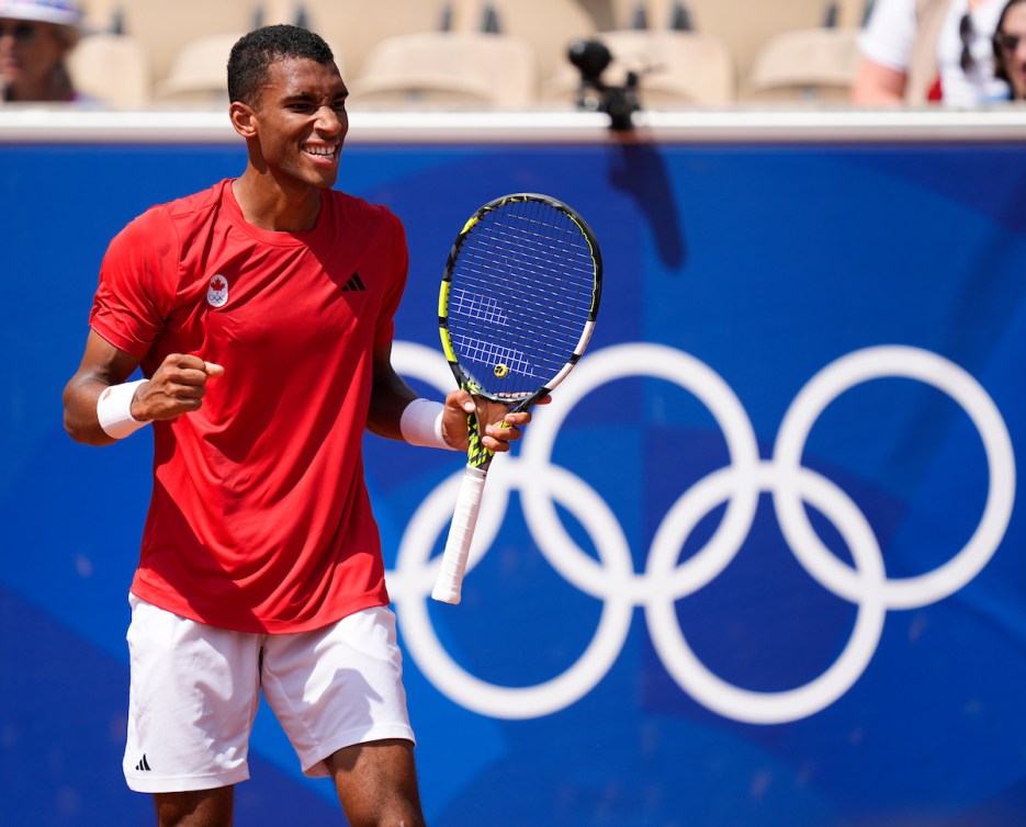 Felix Auger-Aliassime in red shirt and white shorts pumps his fist in front of the Olympic rings