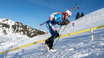 A skier dressed in blue and wearing a helmet walks uphill on snow carrying their skis on their backpack