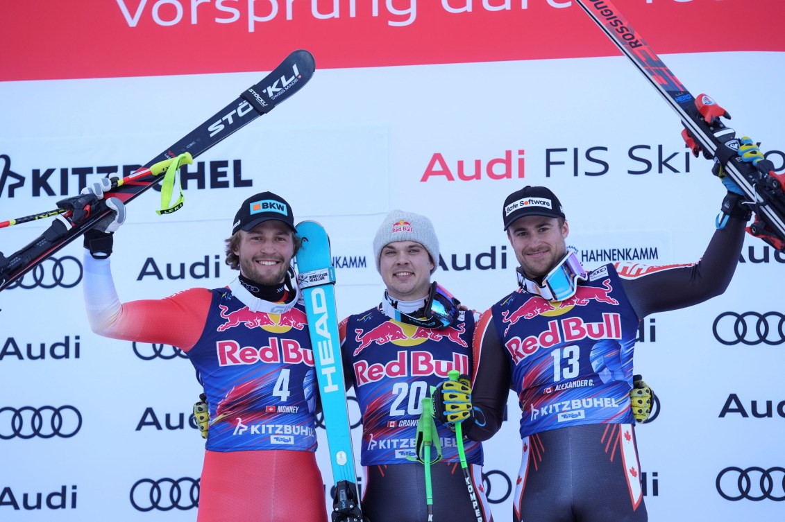 Three male alpine skiers stand in front of the podium backdrop