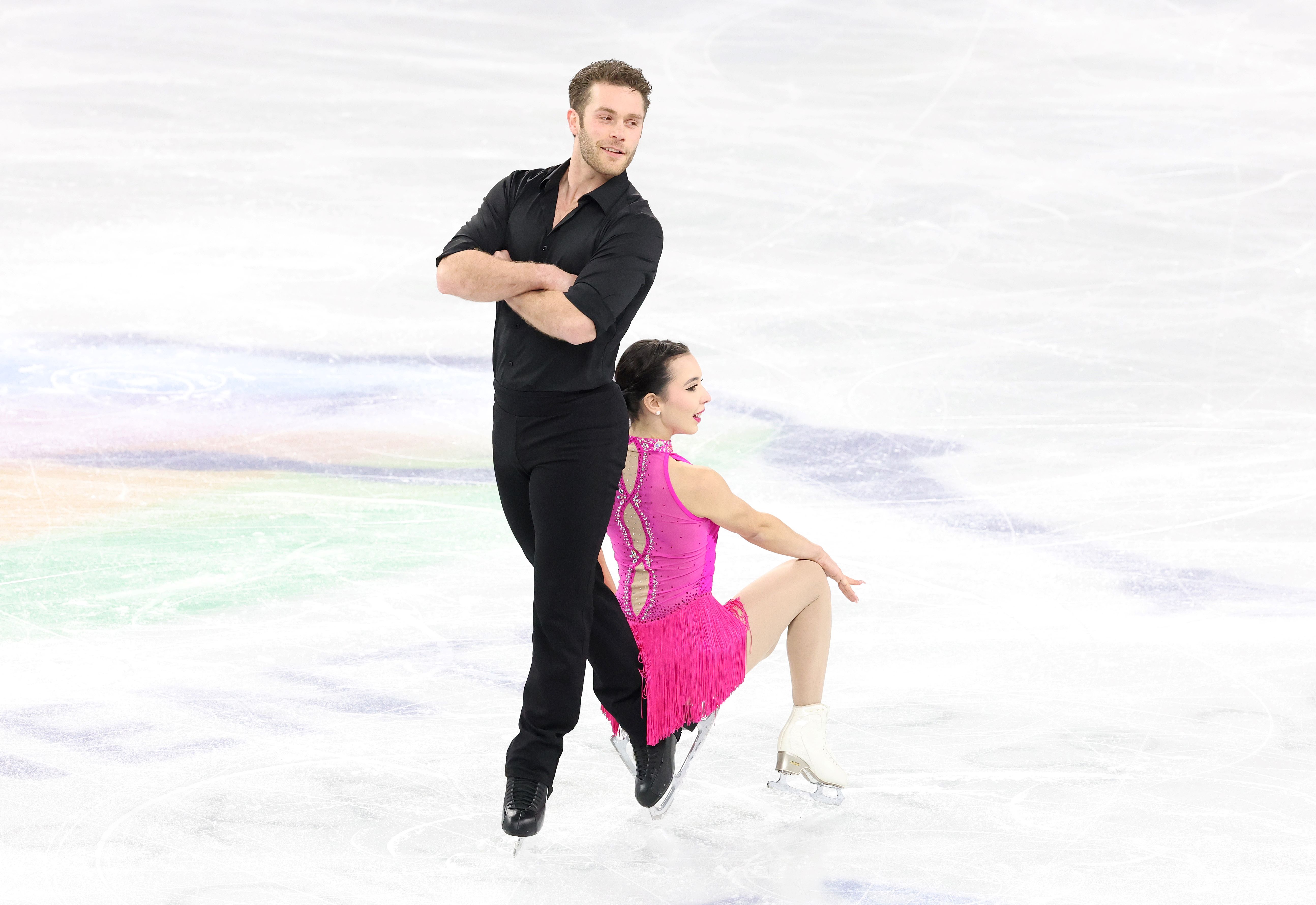 Lia Pereira in a pink dress sits on the skate of partner Trennt Michaud dressed in black pants and shirt