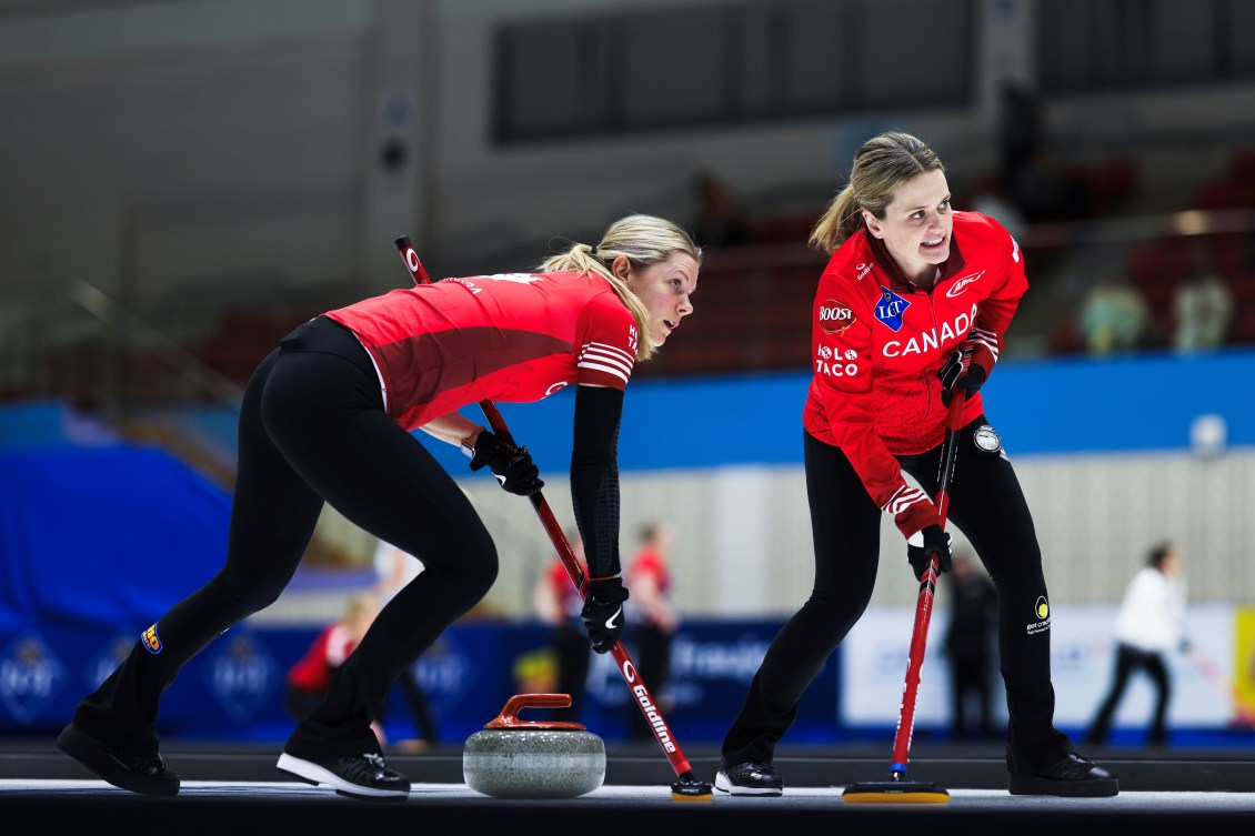 Two female curlers in red shirts and black pants sweep a red stone