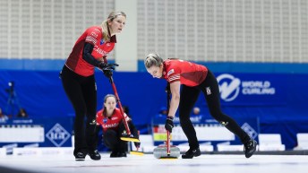 Rachel Homan in a red shirt and black pants watches her stone get swept by her two teammates
