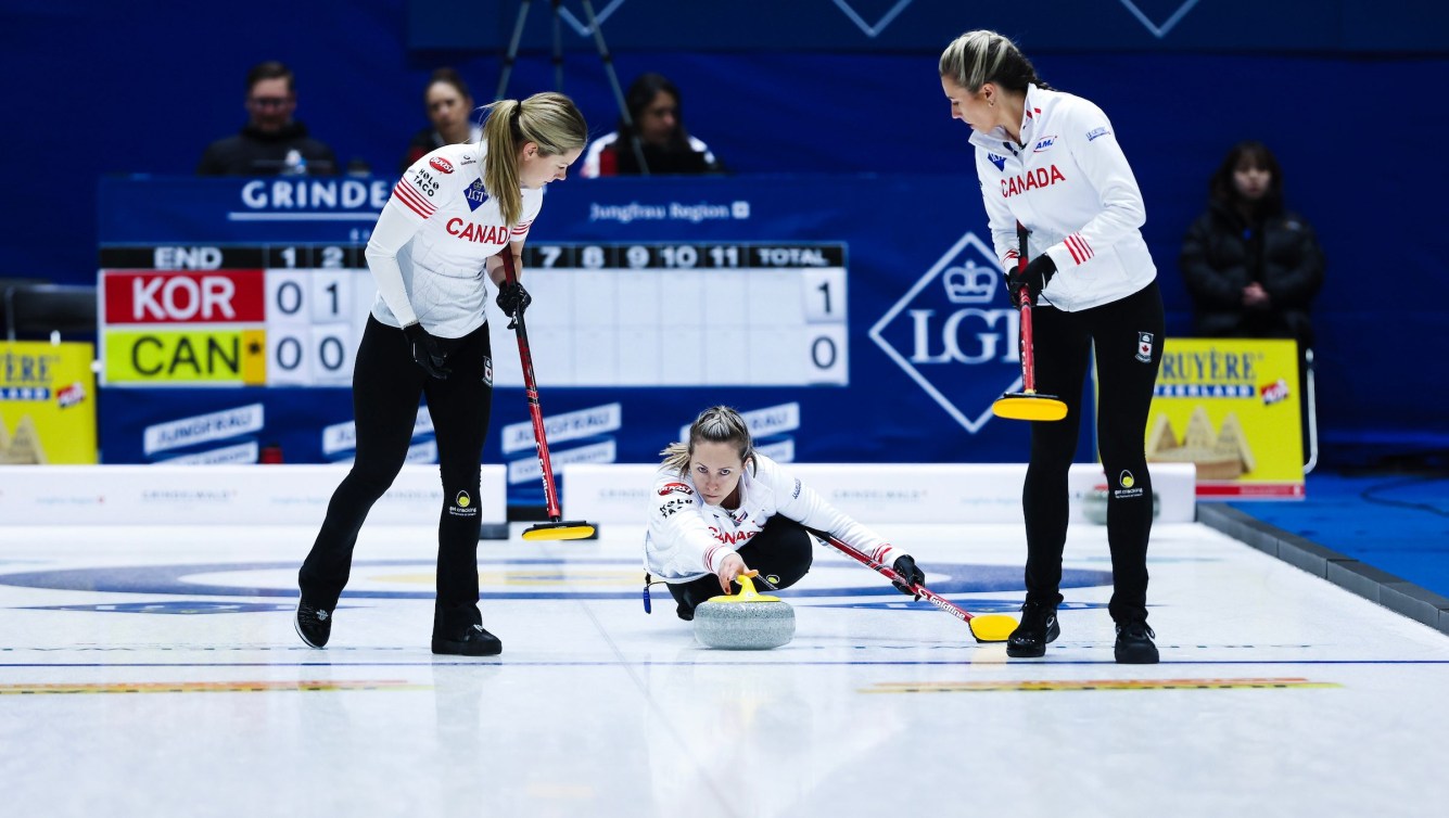Rachel Homan in a white jacket and black pants throws a yellow stone as two teammates prepare to sweep