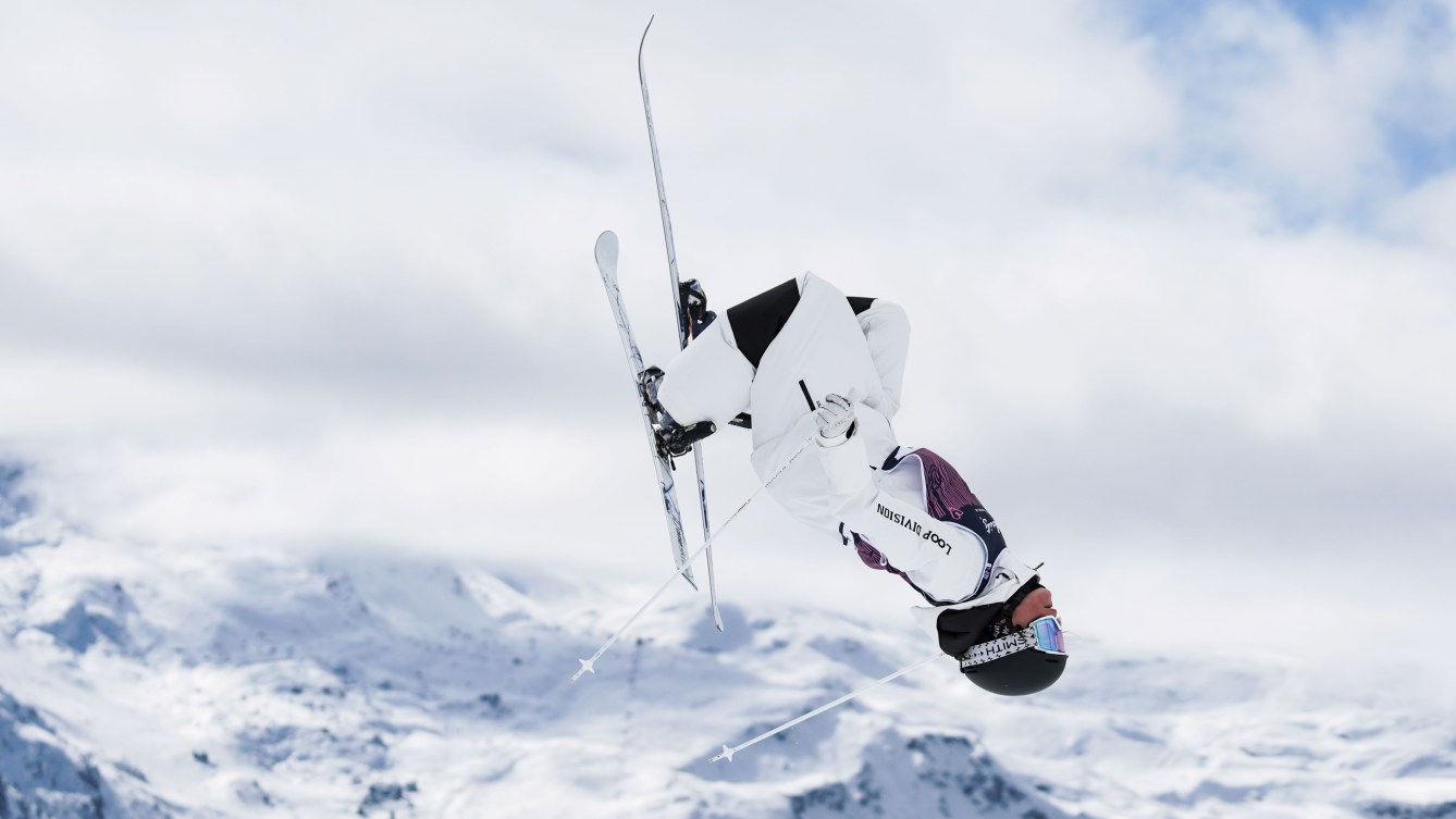 Moguls skier in white snowsuit does a flip in the air with mountains in background
