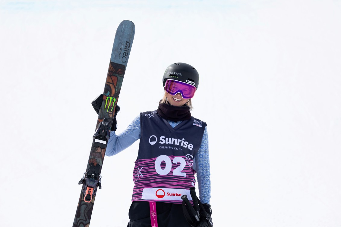 Megan Oldham smiles while holding her skis, wearing a black and purple bib with blue sleeves