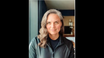 woman with wavy brown hair wearing a black jacket looks straight at camera