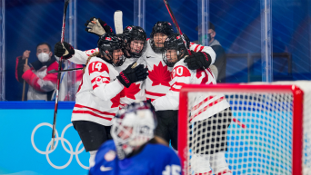 four hockey player's wearing team canada uniforms celebrate a goal behind the team usa goalie's net. they huddle and raise their arms. behind them are blue rink boards with white olympic rings on them