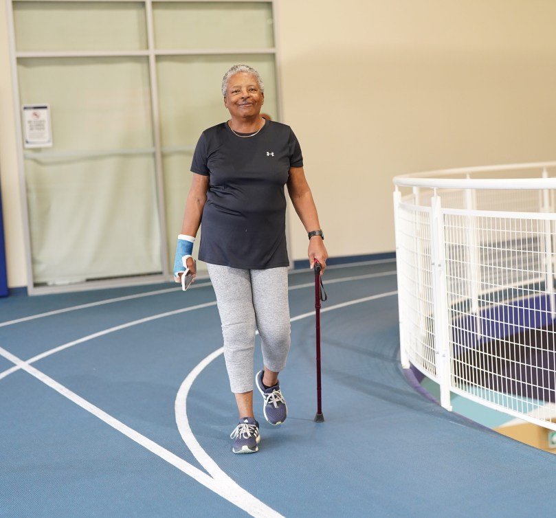 A senior woman in a black shirt and grey pants walks on a blue track with a cane