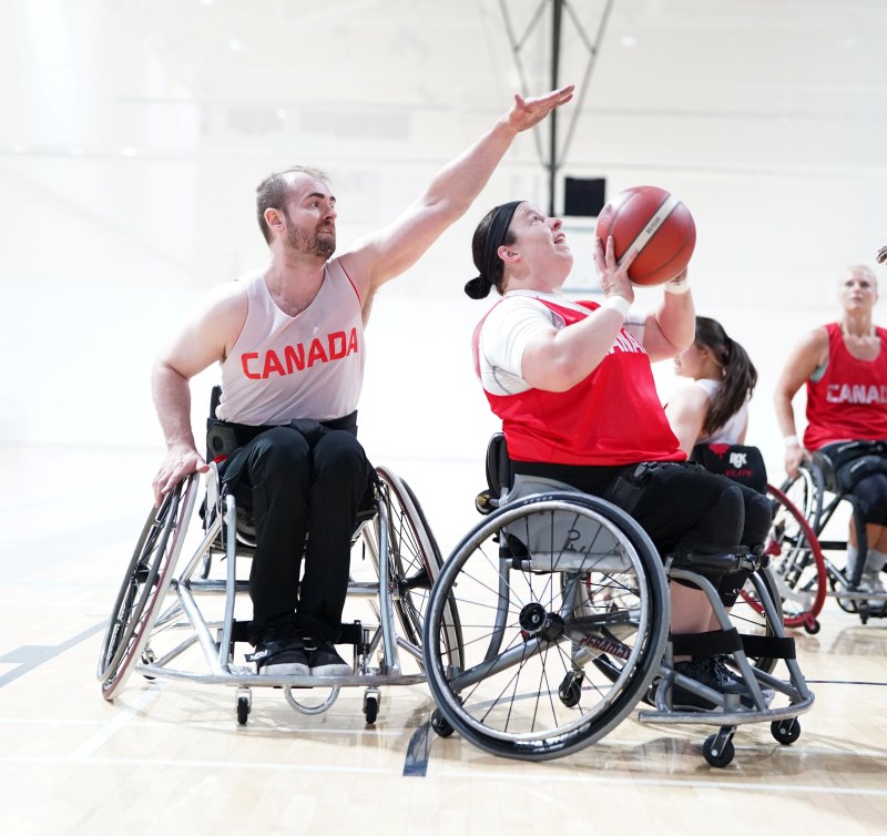 A female wheelchair basketball prepares to shoot the ball while a male player behind her attempts to block