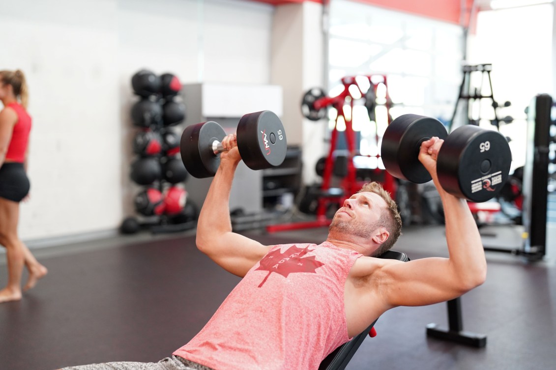 Sam Schachter in a pink tanktop lifts a dumbell in each hand