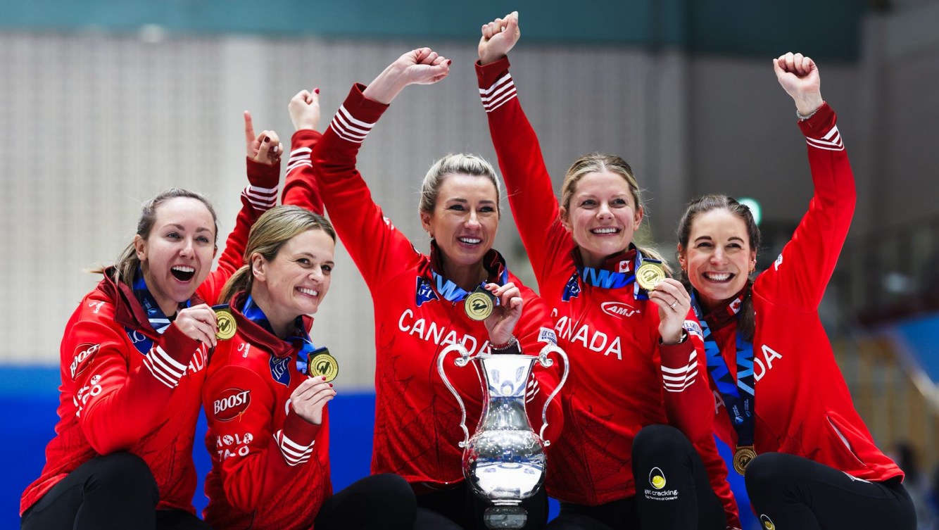 Five women curlers dressed in red jackets celebrate with a trophy while wearing gold medals