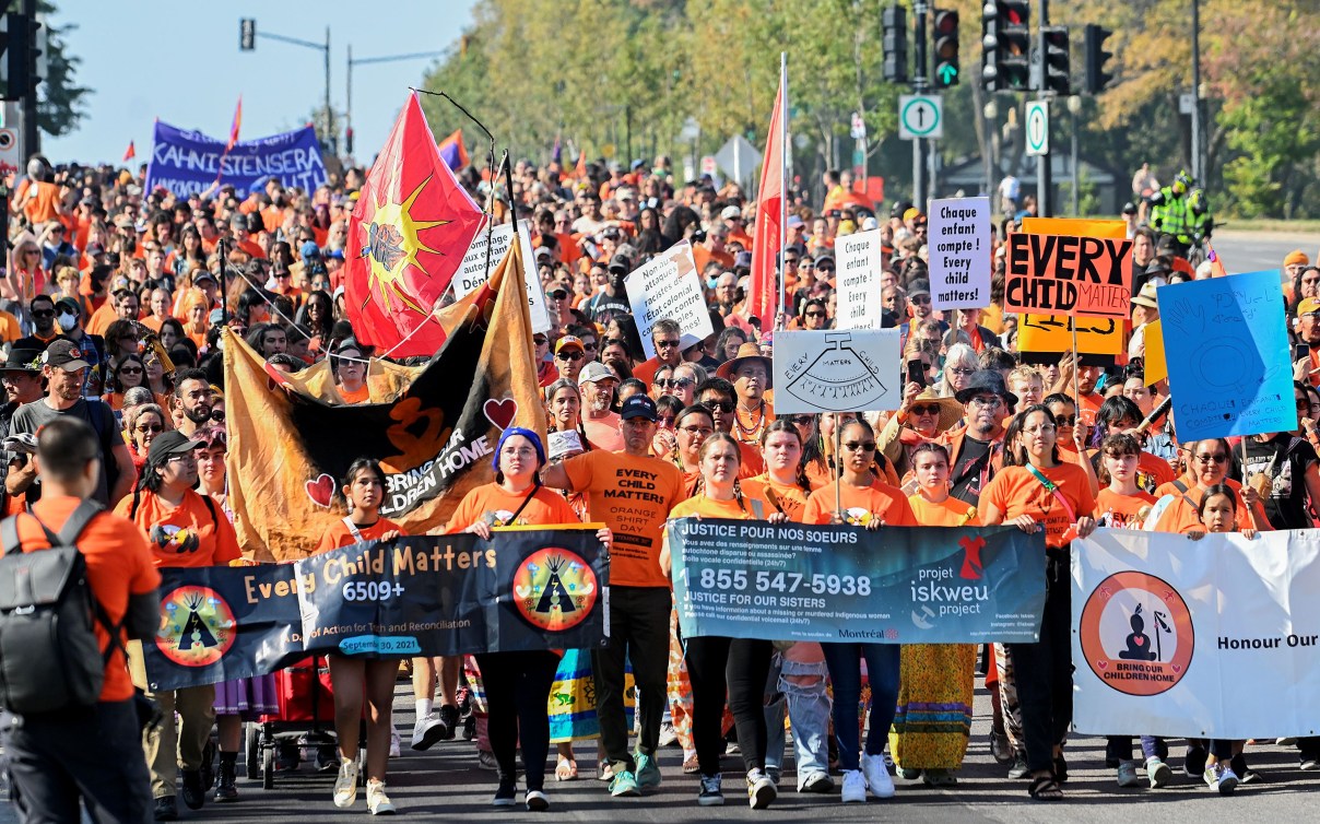 A large mass of people dressed in orange shirts march with signs and flags that say "Every Child Matters"