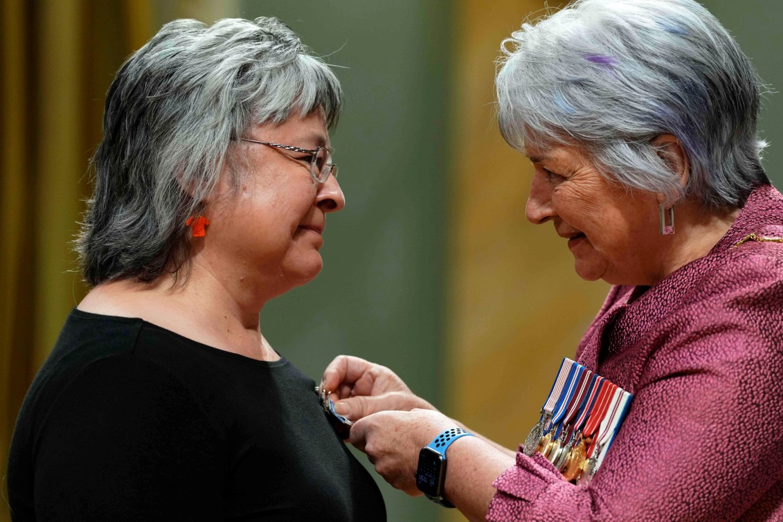 Governor general dressed in pink and wearing medals pins a medal on another grey haired woman in a black shirt