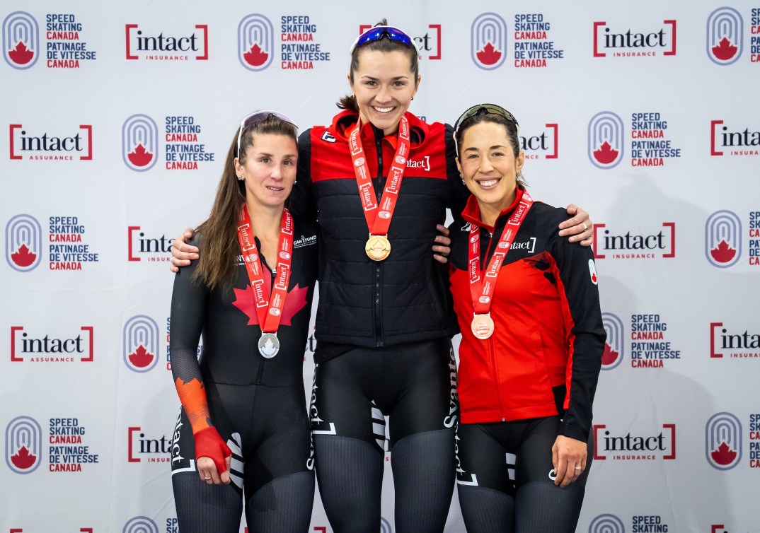 Three female speed skaters in black and red outfits stand with medals in front of a logoed background