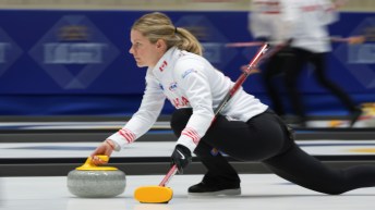 A female curler in white jacket and black pants slides while throwing a stone