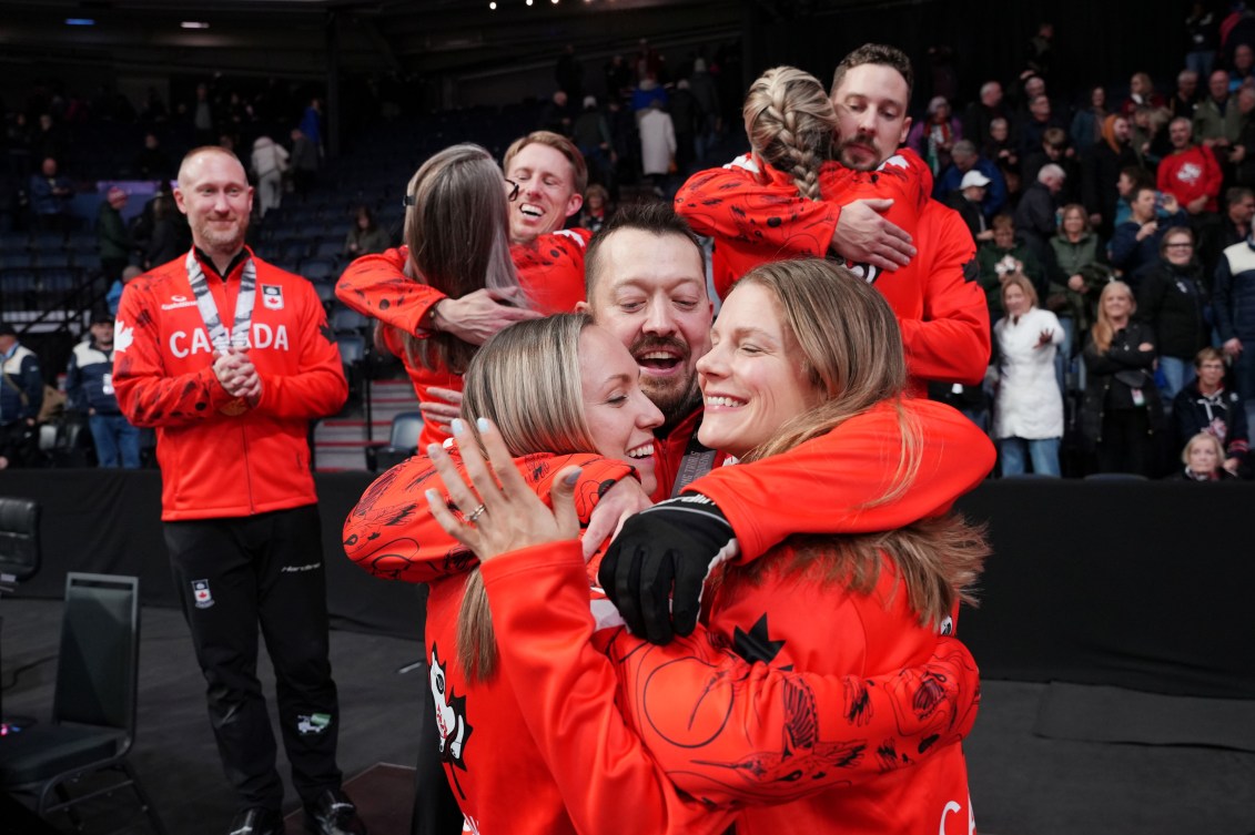 Men's and women's curlers dressed in red jackets hug each other on the podium