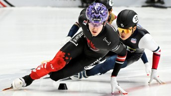 Male short track speed skater in black suit and purple helmet goes around a turn on the ice