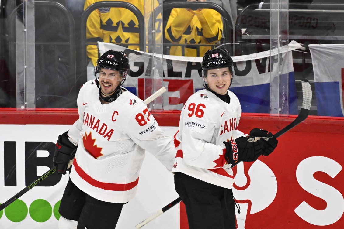Sidney Crosby pats Macklin Celebrini on the back, both players in white jerseys that have a red maple leaf on the front