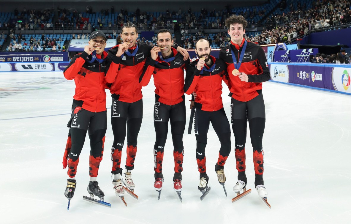 Five male short track speed skaters in red and black suits bite gold medals standing on the ice