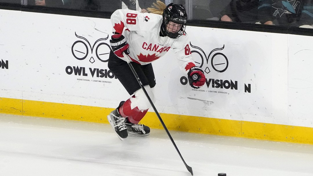 Female hockey player in white jersey with red maple leaf skates along boards with puck on stick