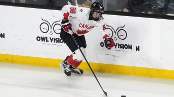 Female hockey player in white jersey with red maple leaf skates along boards with puck on stick