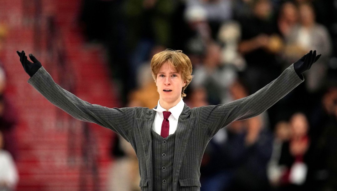 Wearing a grey suit with red tie, blond haired Stephen Gogolev holds his arms out to the side preparing to bow