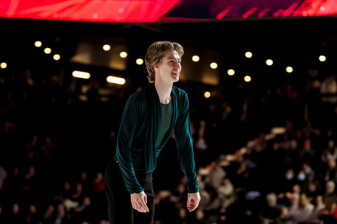 Blond haired man smiles while wearing a forest green shirt in a dark arena