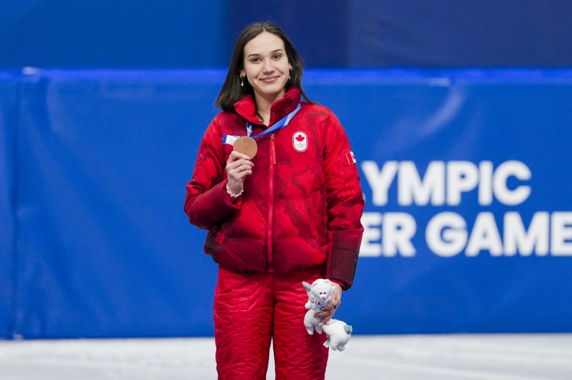 Courtney Sarault celebrates on podium after winning a bronze medal, wearing red Olympic gear.