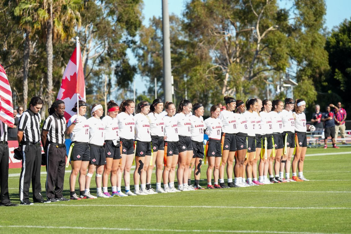 Female flag football players in white shirts and dark shorts stand in a line 