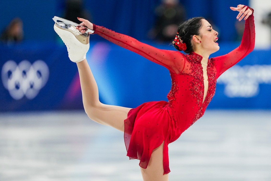 Madeline Schizas in a red long sleeved dress grabs one skate blade with her hand while having her other arm raised